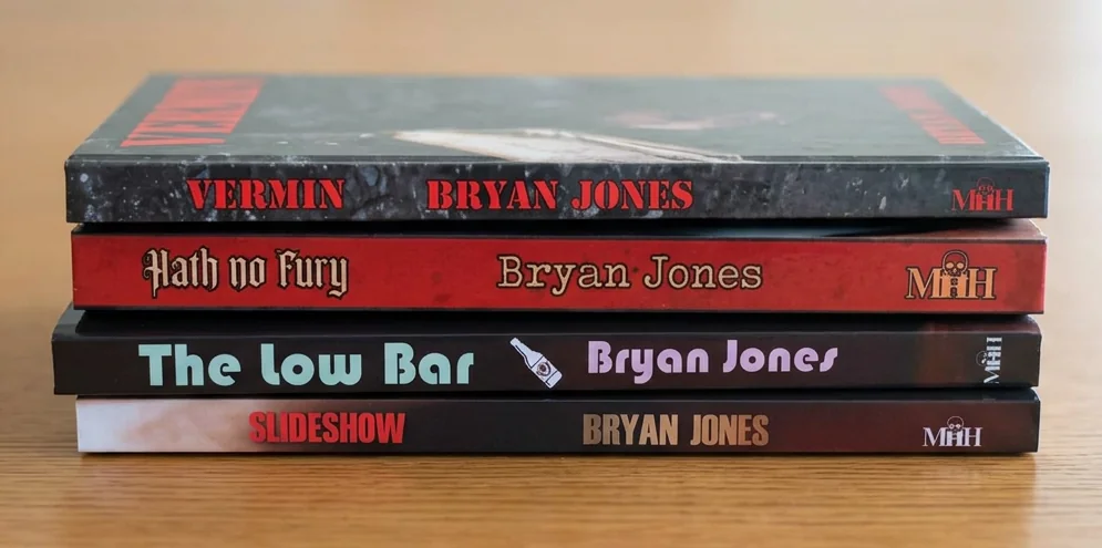 The four Bryan Jones books stacked on a wooden table: Vermin, Hath No Fury, The Low Bar and Slideshow